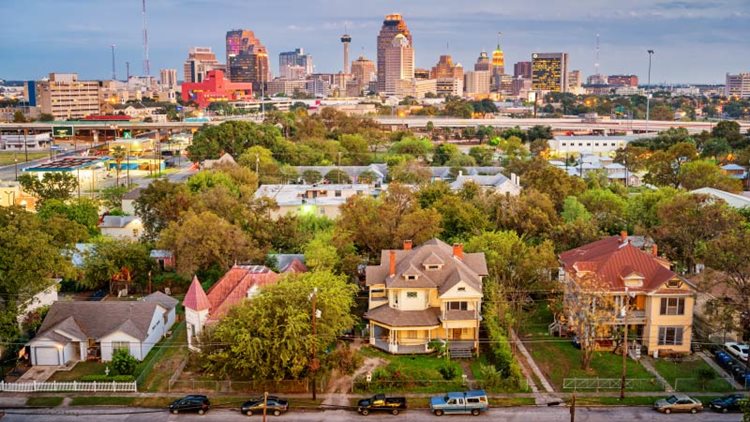 A row of four large residential homes on the outskirts of Downtown San Antonio are facing away from the tall buildings of the city center. Cars and trucks are parked in front of the homes and mature trees are growing in the fronts and backs of the houses.