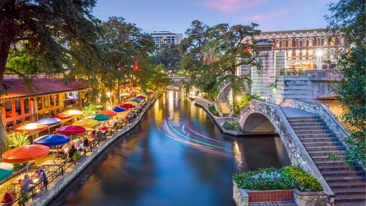 A time-lapse of the River Walk in Downtown San Antonio shows a trail of lights from a boat that just passed under a pedestrian overpass. Lining the river are dozens of bistro tables with colorful umbrellas. Many are occupied with locals and visitors enjoying a beautiful night in San Antonio.