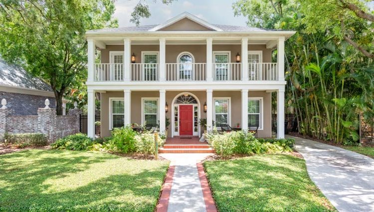 A beautiful two-story, colonial-style home in the Monte Vista neighborhood of San Antonio, Texas. The house features first- and second-story covered porches, lined with traditional pillars. A single gable adorns the roof of the beige home and the front door is painted red as an accent.