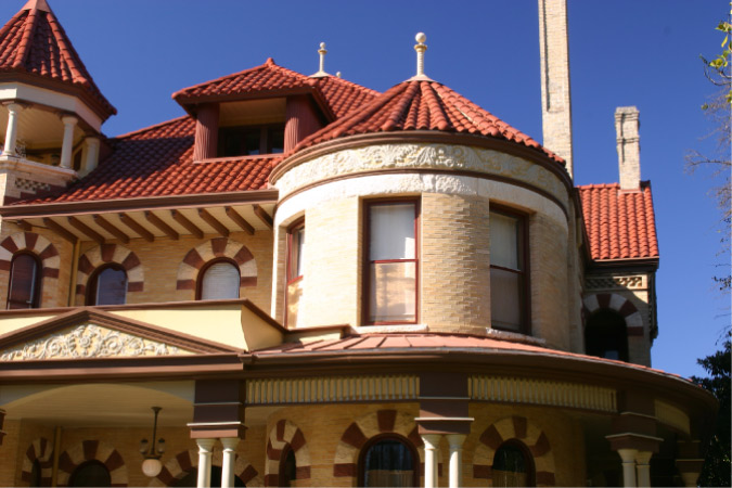 View of the top portion of a beautiful brick and stone home in the King William Historic District of Southtown, a neighborhood in San Antonio. The home is styled like a Mediterranean castle, with clay roof tiles, rounded windows, and a stout tower on the second floor.