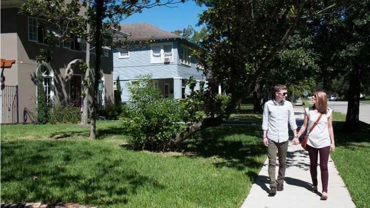 A happy young couple is holding hands and taking a walk through the Alamo Heights neighborhood in San Antonio. It’s a sunny day, so they’re both wearing sunglasses. The man is looking at the woman and the woman is looking at the large, historic homes in the neighborhood.