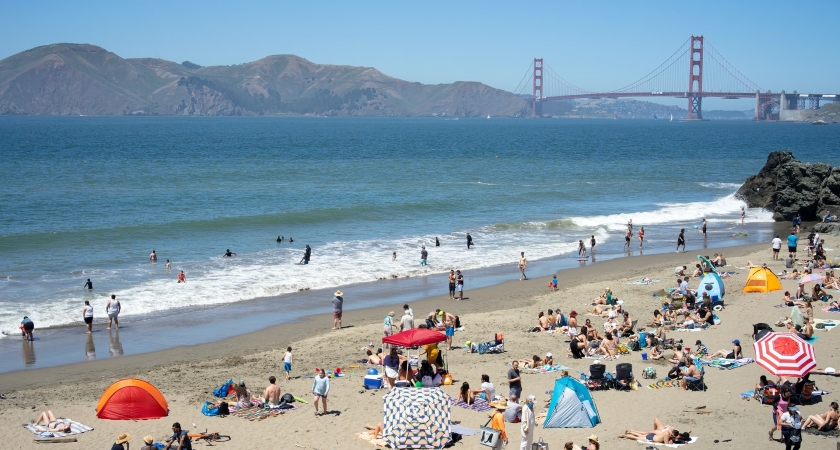 Dozens of beachgoers enjoy a sunny day frolicking on a San Francisco beach. The Golden Gate Bridge and nearby mountains are visible in the distance. 