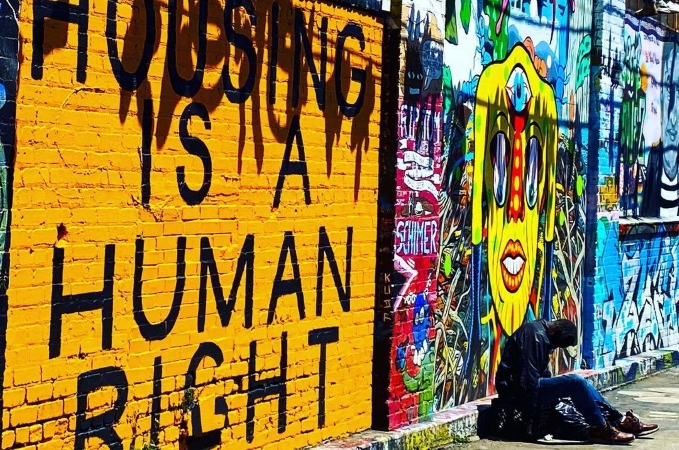 A homeless man is sitting against a colorfully muraled wall in San Francisco. The brick wall beside him has the message “HOUSING IS A HUMAN RIGHT” painted in large black letters against a bright yellow background.