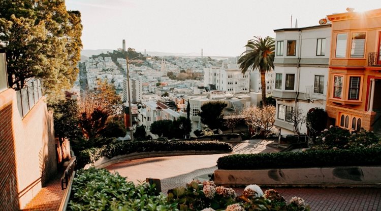 A view down winding Lombard Street in San Francisco, California, in the early morning. The city and one of its bridges can be seen below in the distance. 