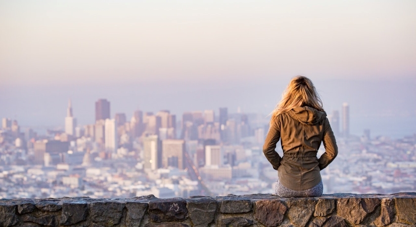 A young woman is sitting on a stone wall with her hands in her coat pockets, looking out at the San Francisco skyline in the distance.