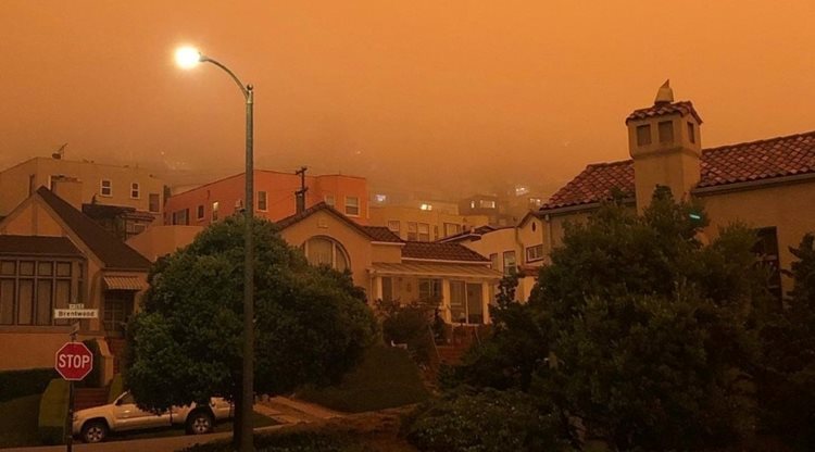 A San Franciscan neighborhood is filled with smoke from wildfires. The whole scene has an orange glow to it, and though it’s still daytime, the street lights have come on due to the limited natural light.