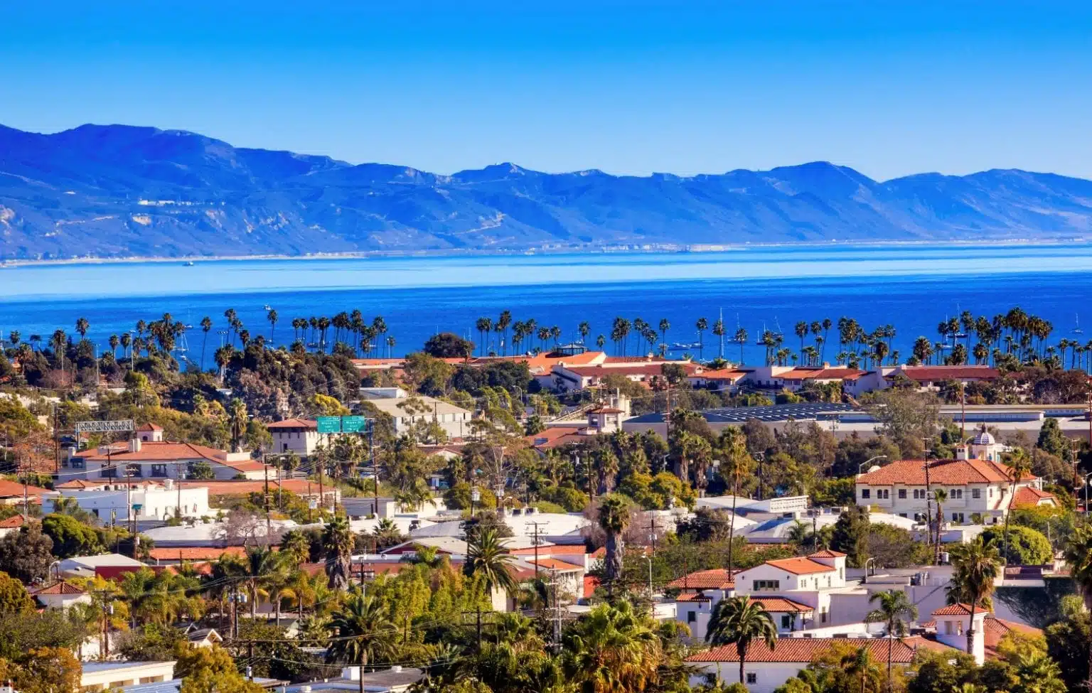 View of the ocean and mountains in Santa Barbara, California, from across the tops of palm trees and the red tile roofs of the city buildings