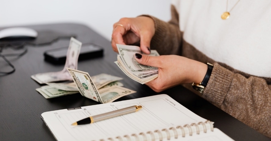 Close-up view of a woman counting money at her desk. She has a notepad and pen in front of her for keeping notes.