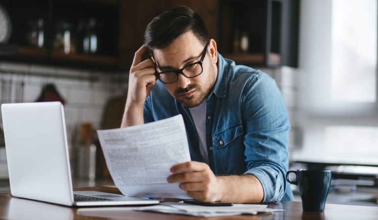 A man is sitting in his kitchen with his laptop in front of him. He’s holding some financial documents and looking over his property taxes. He seems stressed. 