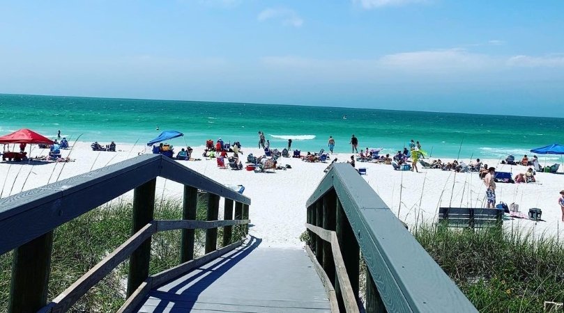 View of Pass-A-Grille Beach and the water from a wooden pathway. Dozens of people are relaxing in the sand under umbrellas, enjoying a perfect summer day.
