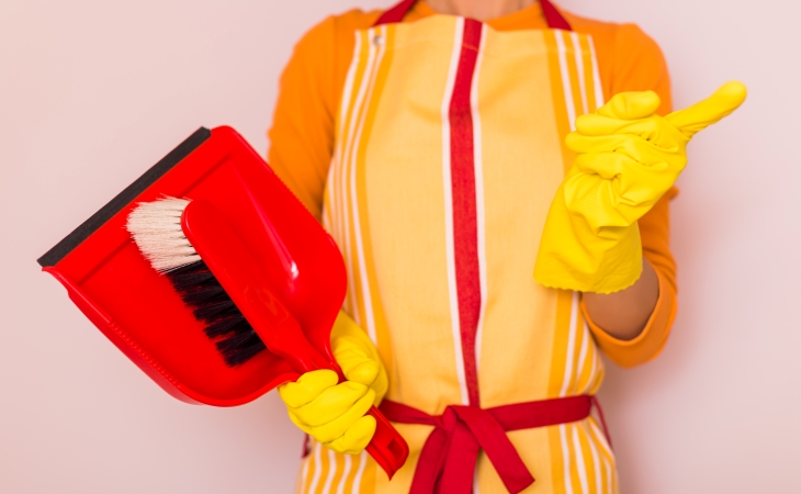 A close-up view of the torso of a woman. She is wearing a yellow and red cleaning apron and yellow rubber gloves. She’s holding a handheld broom and dustpan in one hand and is waving a finger with the other. 