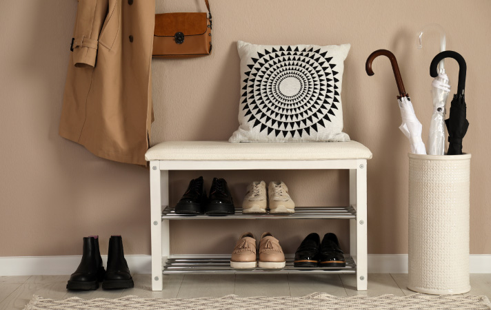 A stylish entryway highlighting a bench that does double-duty as a shoe rack, along with a coat on a coat rack, umbrellas in an umbrella storage container, and a pillow on top of the bench.