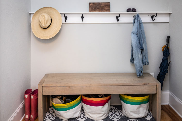 View of one side of a mudroom, featuring hooks to hang coats and hats, a bench, and three baskets used for storing shoes.