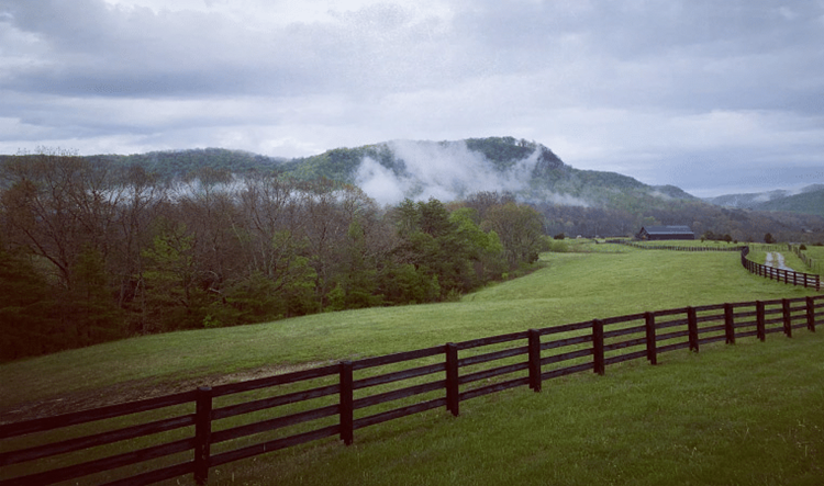 The forests and mountains around Berea, Kentucky, in the early morning. A misty fog is approaching beneath a cloudy sky. In the foreground are mowed fields and a long, wooden fence, and in the distance is a large barn. 