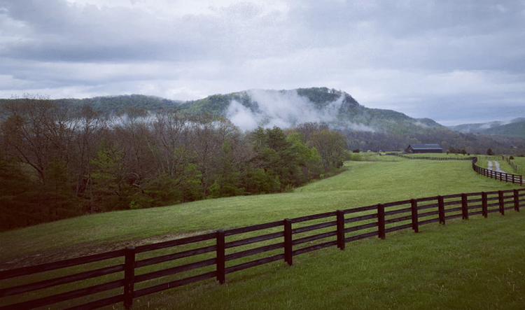 The forests and mountains around Berea, Kentucky, in the early morning. A misty fog is approaching beneath a cloudy sky. In the foreground are mowed fields and a long, wooden fence, and in the distance is a large barn.