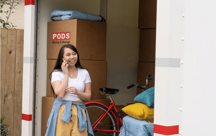 A young woman is smiling as she takes a phone call by the entrance to her PODS portable moving container. The container is partially loaded with moving boxes, pillows, and a bike. 