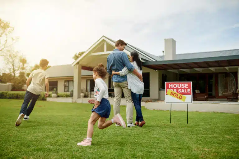 A husband and wife embrace in front of their newly purchased home as their two kids play in the yard.