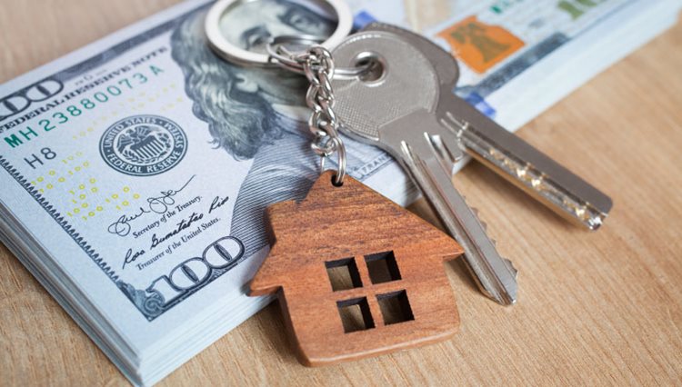 A set of keys with a wooden keychain in the shape of a house is sitting on top of a stack of 100 dollar bills, representing the concept of a down payment on a house.