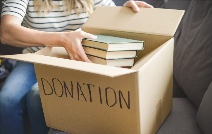 A woman is sitting on her couch, placing books in a box labeled “DONATION.”