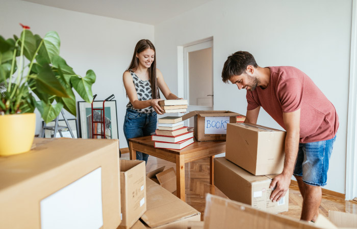 A young couple is in the process of moving house. The man is picking up two moving boxes and the woman is packing books into another box.