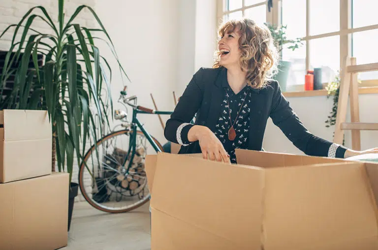 A woman is smiling as she packs up a moving box in her living room. There’s a houseplant, a bicycle, and two other moving boxes in the room beside her.