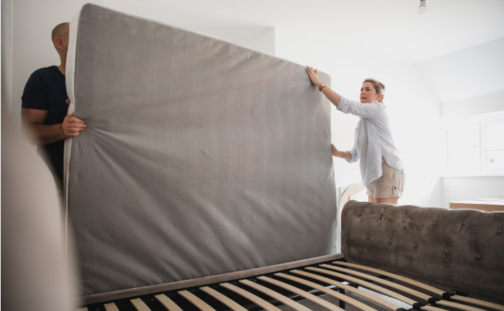 A couple works together to move a mattress off of a bed. They’re preparing to pack it for a move.