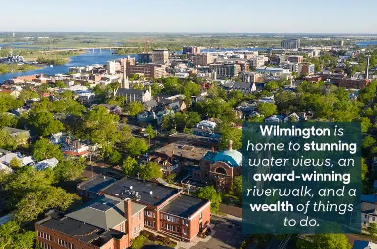Springtime aerial view of downtown Wilmington, NC. The overlaid text reads “Wilmington is home to stunning water views, an award-winning riverwalk, and a wealth of things to do.”