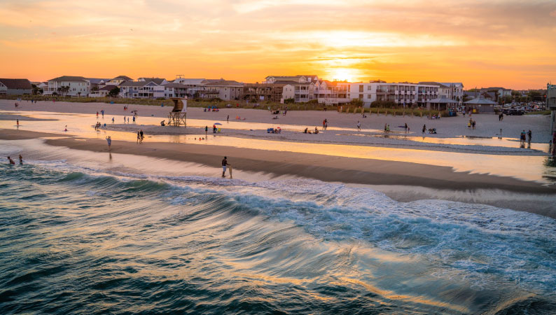 Sunset at Wrightsville Beach in North Carolina. Dozens of people walk along the sand as the sky turns a peachy orange. 