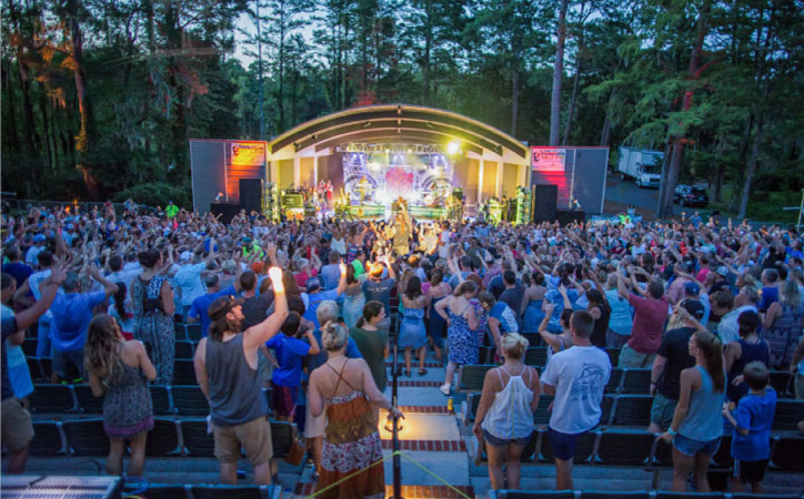 A live show at Greenfield Lake Amphitheater has filled the seats with spectators. The lights from the stage contrast beautifully as the sun’s light fades behind the trees.