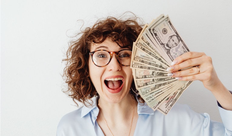 A young woman smiles a cheesy grin as she fans a handful of bills in front of her face.  (Source: Karolina Grabowska via Pexels)