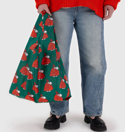 Close-up of the bottom half of a person in jeans and a sweater, holding a holiday-themed reusable bag from Baggu