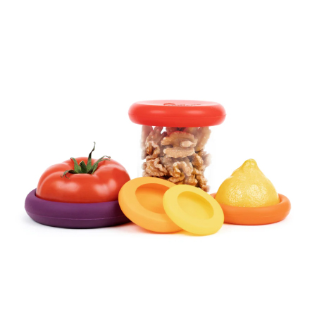 Food huggers being used to keep a half of a tomato, half of a lemon, and an opened jar of walnuts fresh