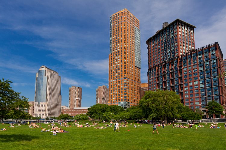 Dozens of locals are enjoying a sunny day outdoors in Battery Park City, New York, with tall residential buildings rising up in the background. 