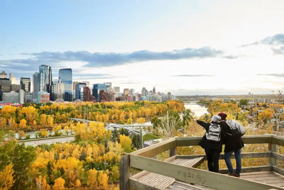 Two day-hikers looking out across a river at Downtown Calgary and into distant Calgary neighbourhoods. They’re standing on a wooden platform above a lush autumnal forest.