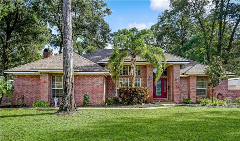 A one-story brick home in the Holly Oaks neighborhood of Jacksonville, FL. It has a large green lawn and is surrounded by various types of trees, from pines to palms.
