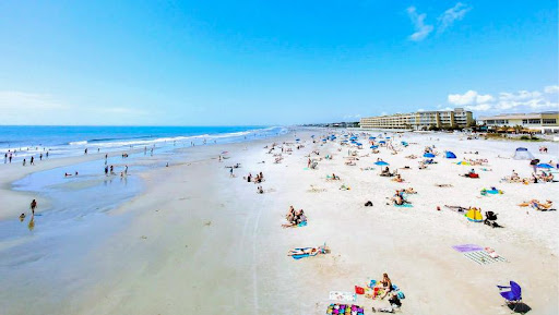 Beachgoers are seen relaxing on the sand and playing in the water at Folly Beach, SC. High-rise condominiums can be seen in the distance.