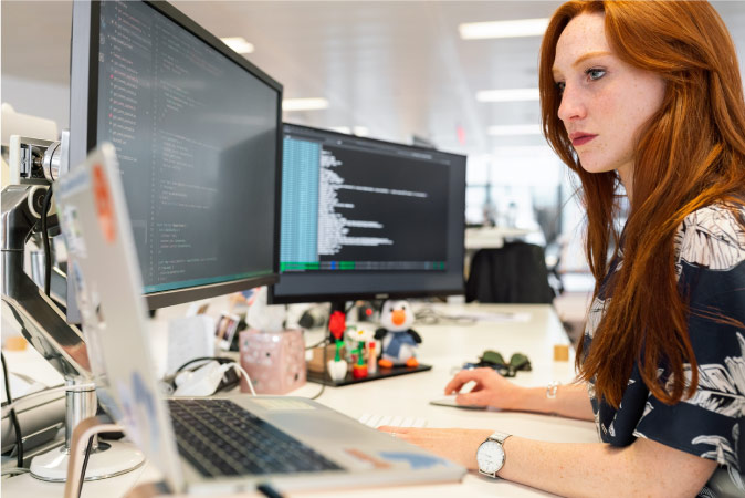A woman with long red hair is working at a desk with three monitors in Charleston, SC.