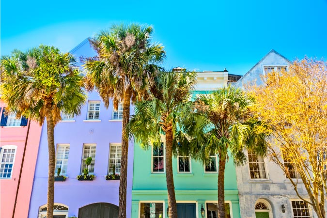 A street-level view of palm trees in front of a row of colorful townhomes on Rainbow Row in Charleston, South Carolina.