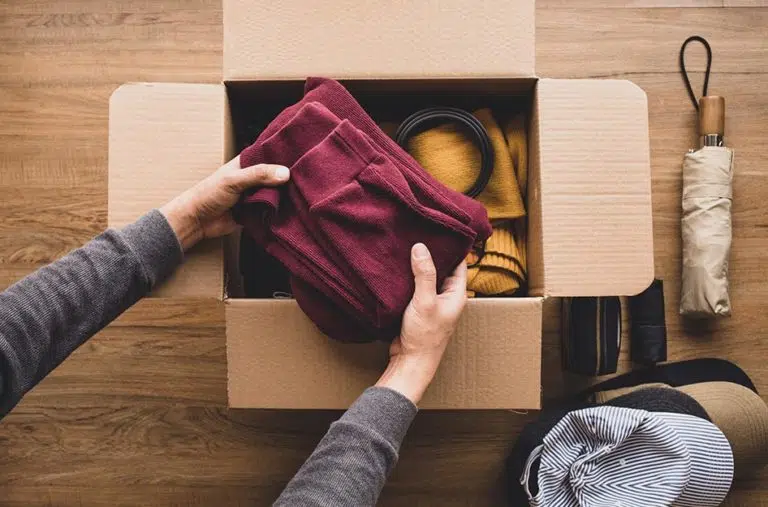 View from above of a person packing nicely folded clothes into a moving box. There is a pile of hats and a closed umbrella nearby.
