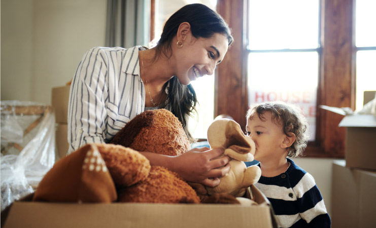 A happy young mother packs stuffed animals away in moving boxes as her toddler son looks on.
