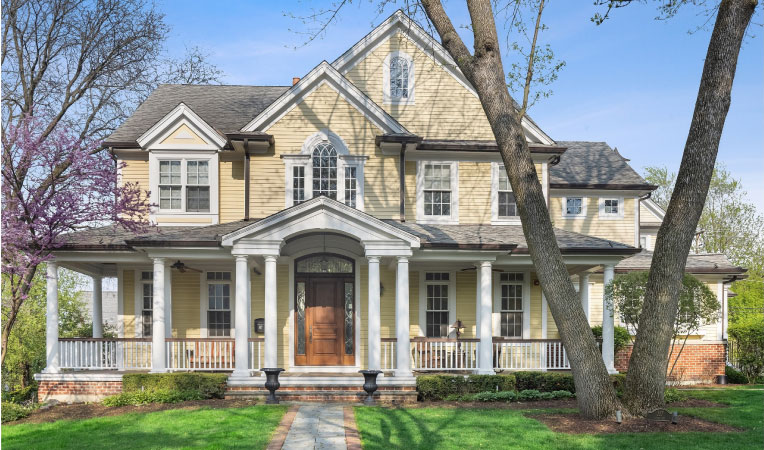 A two-story residential home in Chicago’s Clarendon Hills suburb. The home has a wood siding exterior, painted a light yellow with white trim. There are over ten ionic columns around the covered porch and entrance to the home. The lawn is a lush green and there’s a mature tree in the front yard.