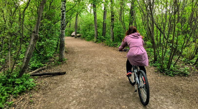 A woman in a purple coat is riding her bike on a wooded trail in Edmonton, Alberta.
