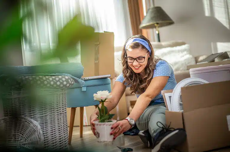 A young woman is sitting on the floor among several packed moving boxes. She’s only just started unpacking and appears excited as she places a blooming houseplant on the floor of her new home.