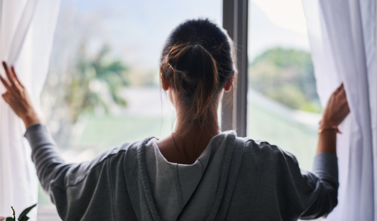 A young woman is holding white curtains open as she admires the view from the large windows of her new home.