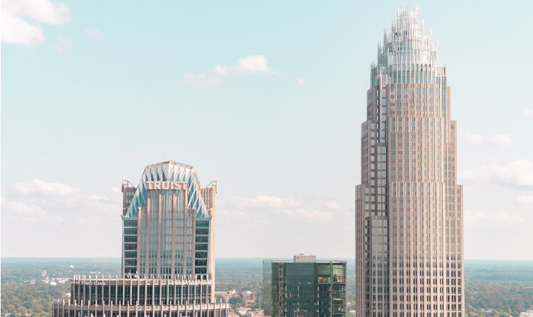 A rooftop view of two skyscrapers in Charlotte, NC, including the Bank of America Tower. In the distance, Charlotte and neighboring suburbs sprawl out into the horizon.