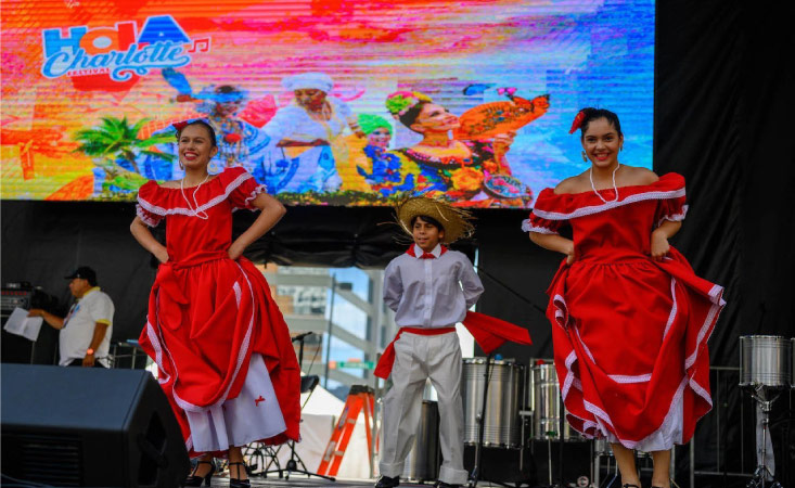 Hispanic dancers are performing on a stage in front of a screen that says “Hola Charlotte” during one of the many festivals the city offers.