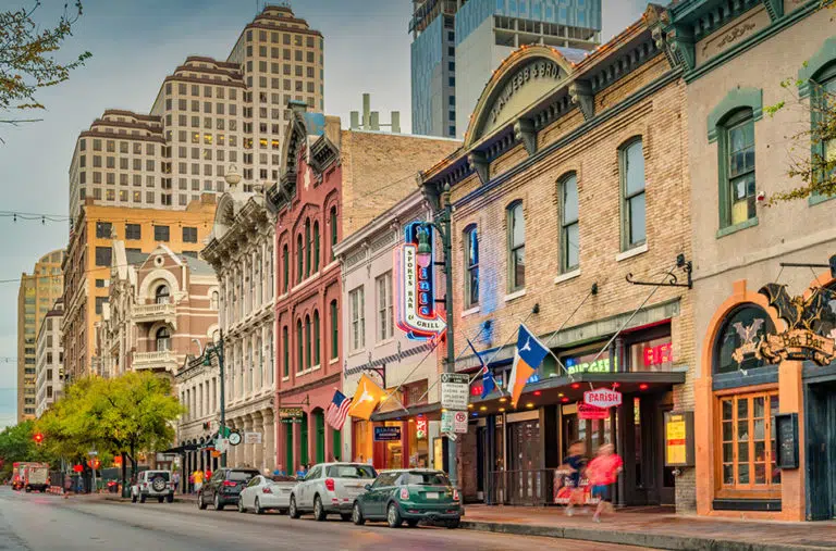 A portion of the Sixth Street music and entertainment district in Austin, Texas, on a clear afternoon. One of the storefronts features a large overhang with various flags posted from it.