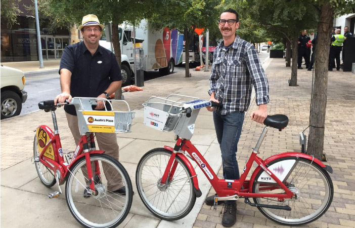 Two smiling men stand beside their bike rentals in Austin, Texas. The bikes are both red and have baskets fitted to their handlebars.