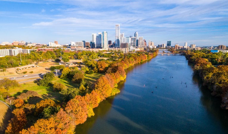 Aerial view of Lady Bird Lake in Austin, Texas, in the fall. The trees that line the waterway have begun to change colors, and the Austin skyline is visible in the distance. 
