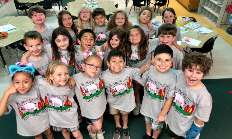 A group of first graders from Austin ISD smile for a picture in their classroom. The kids are all wearing matching gray shirts with a cartoon graphic on the front.